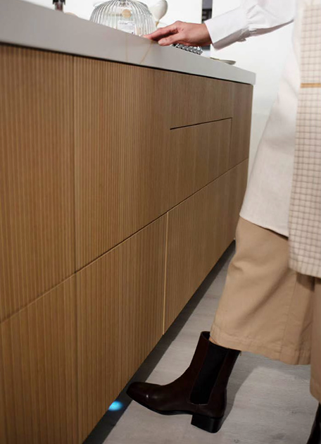 Mueble de cocina con diseño elegante en madera estriada, que incorpora tecnología táctil con sensor de pie iluminado, permitiendo abrir cajones sin manos. Funcionalidad y estilo se unen en un gesto práctico y moderno.