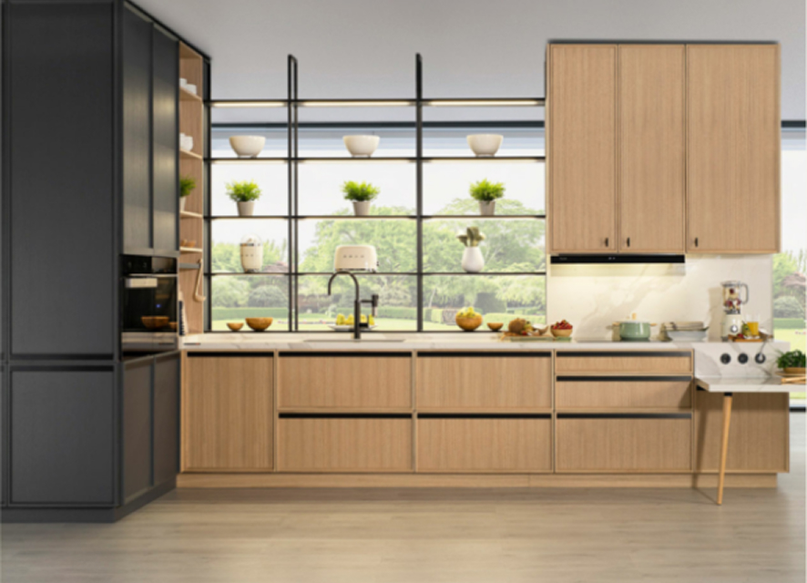 Cocina de estilo moderno con muebles de líneas limpias en acabado madera clara. Un módulo alto negro alberga electrodomésticos integrados, mientras que la isla se extiende con una mesa auxiliar tipo barra. Estanterías abiertas frente a ventanales amplios aportan luz natural y un toque decorativo con plantas y vajilla.