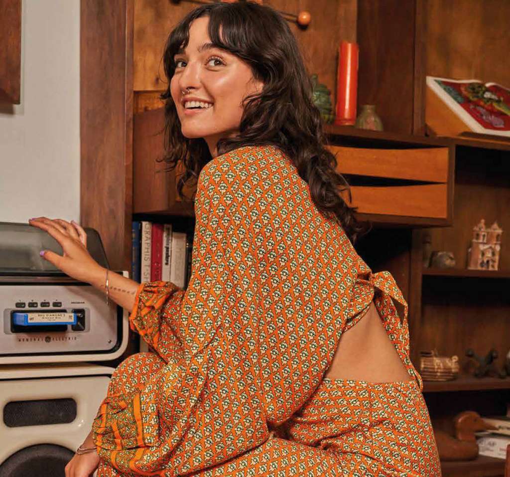 Una chica onríe junto a un tocadiscos vintage, vestida con un conjunto estampado que evoca libertad y estilo retro. El ambiente cálido y lleno de detalles habla de historias, música y alma.