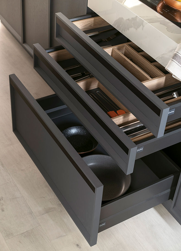 tres cajones abiertos de un mueble de cocina en acabado oscuro. En el cajón superior hay un organizador de madera con compartimentos para cuchillos y utensilios, el cajón intermedio contiene divisores para cubiertos y herramientas de cocina, y el cajón inferior almacena sartenes y planchas. El interior en tono madera clara contrasta con el frente oscuro, ofreciendo una organización clara y accesible.