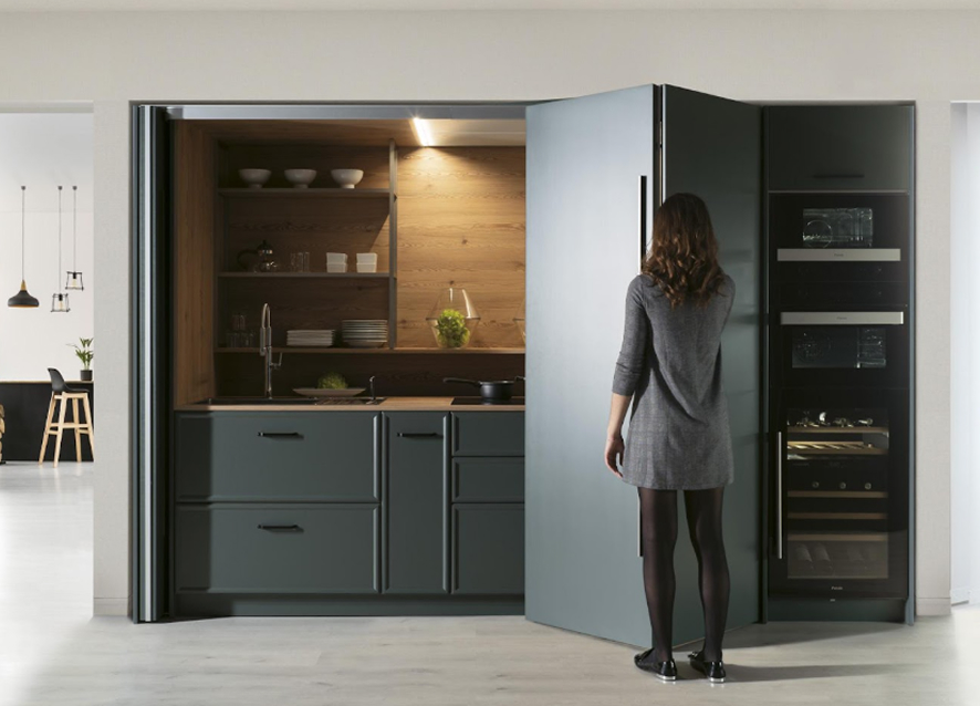 Cocina compacta y funcional con frentes en gris grafito mate y tiradores integrados. El interior, revestido en madera natural, aporta calidez y contraste. La iluminación led bajo estante resalta el fondo y crea una atmósfera acogedora. Las puertas escamoteables permiten ocultarla por completo, ideal para espacios abiertos o multifuncionales.