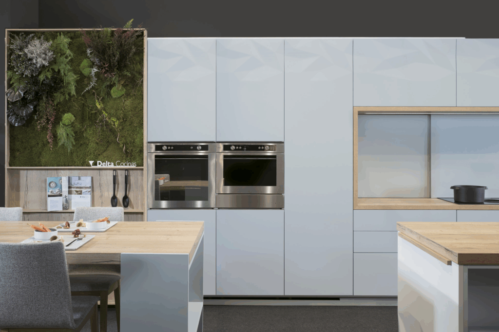 Frentes de armario en acabado blanco mate con textura facetada, integrados con electrodomésticos en acero inoxidable. Panelado continuo con nicho de trabajo enmarcado en madera clara. Diseño pulcro y elegante.