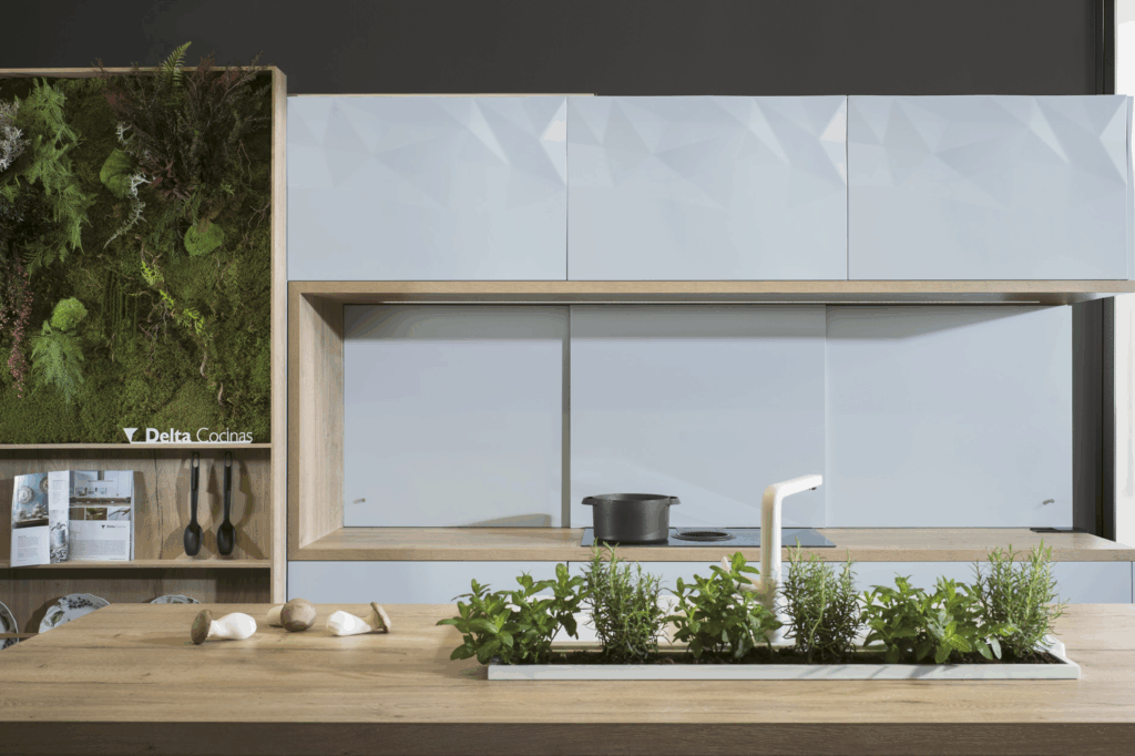 cocina con mobiliario de líneas rectas y estilo moderno. Los muebles altos tienen puertas lisas en acabado blanco con un sutil relieve geométrico. En la parte inferior, una estructura de madera enmarca la zona de cocción, que incluye puertas correderas. La isla central combina madera clara en la encimera y módulos blancos en la parte inferior, algunos con estantes abiertos. También se puede ver una estantería vertical abierta en uno de los laterales, y un panel decorativo con vegetación sobre una estructura de madera.
