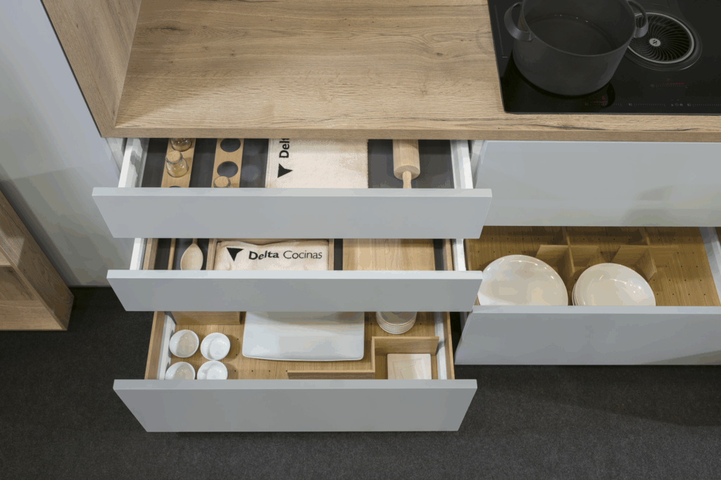 Cajoneras inferiores con frentes en blanco mate, integradas bajo una encimera de madera clara. El sistema de organización interior optimiza el almacenamiento de utensilios, vajilla y accesorios de cocina. Diseño práctico y elegante que combina funcionalidad y estética.