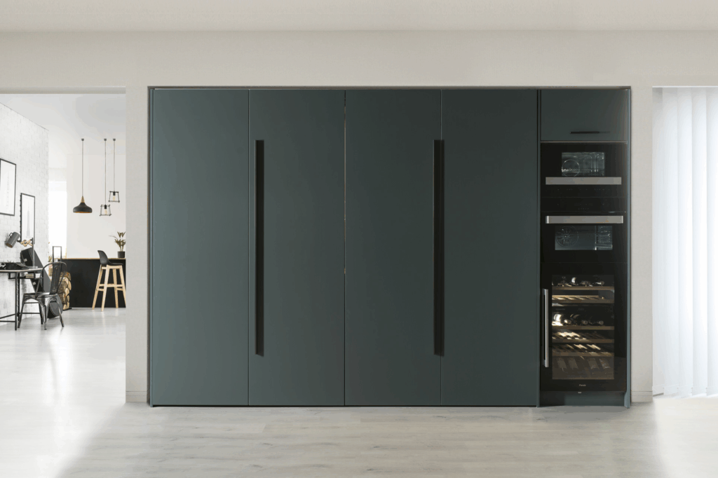 Cocina oculta tras elegantes puertas en gris oscuro con tiradores integrados en negro, que aportan sobriedad y limpieza visual. A la derecha, una columna de electrodomésticos empotrados remata el conjunto con funcionalidad y diseño.