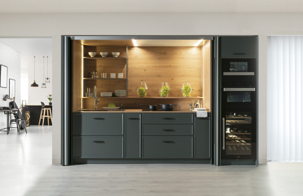 Cocina compacta y funcional con frentes en gris grafito mate y tiradores integrados. El interior, revestido en madera natural, aporta calidez y contraste. La iluminación led bajo estante resalta el fondo y crea una atmósfera acogedora. Las puertas escamoteables permiten ocultarla por completo, ideal para espacios abiertos o multifuncionales.
