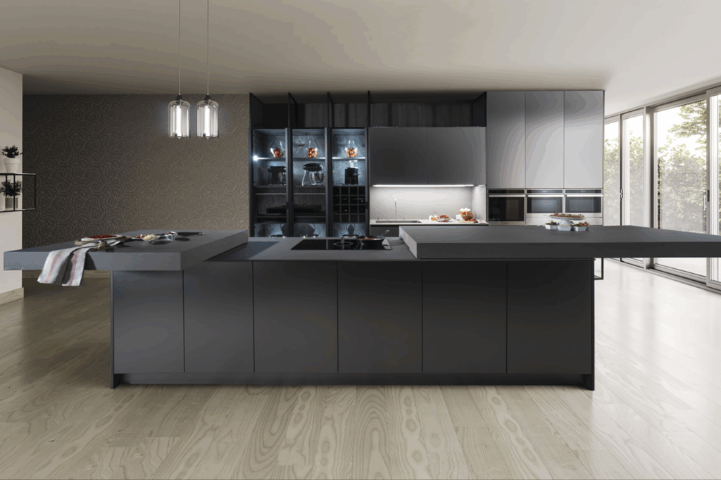 Cocina de diseño vanguardista con mobiliario en tonos gris mate y antracita, sin tiradores, que refuerza la estética minimalista. Una gran isla central con volúmenes desfasados y superficies amplias funciona como área de trabajo y barra. Al fondo, columnas de almacenamiento integran hornos en acero y vitrinas iluminadas, creando un equilibrio entre funcionalidad y exposición elegante. El conjunto se completa con muebles altos en acabados suaves y continuos, resaltando la armonía y sofisticación del espacio.