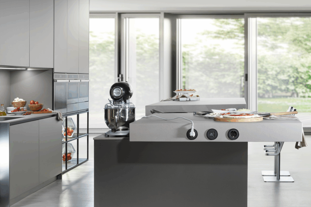 Isla multifuncional con superficie desfasada y enchufes integrados, ideal para cocinar y socializar. Muebles columna en acabado gris suave con vitrina inferior iluminada, combinan elegancia y funcionalidad.