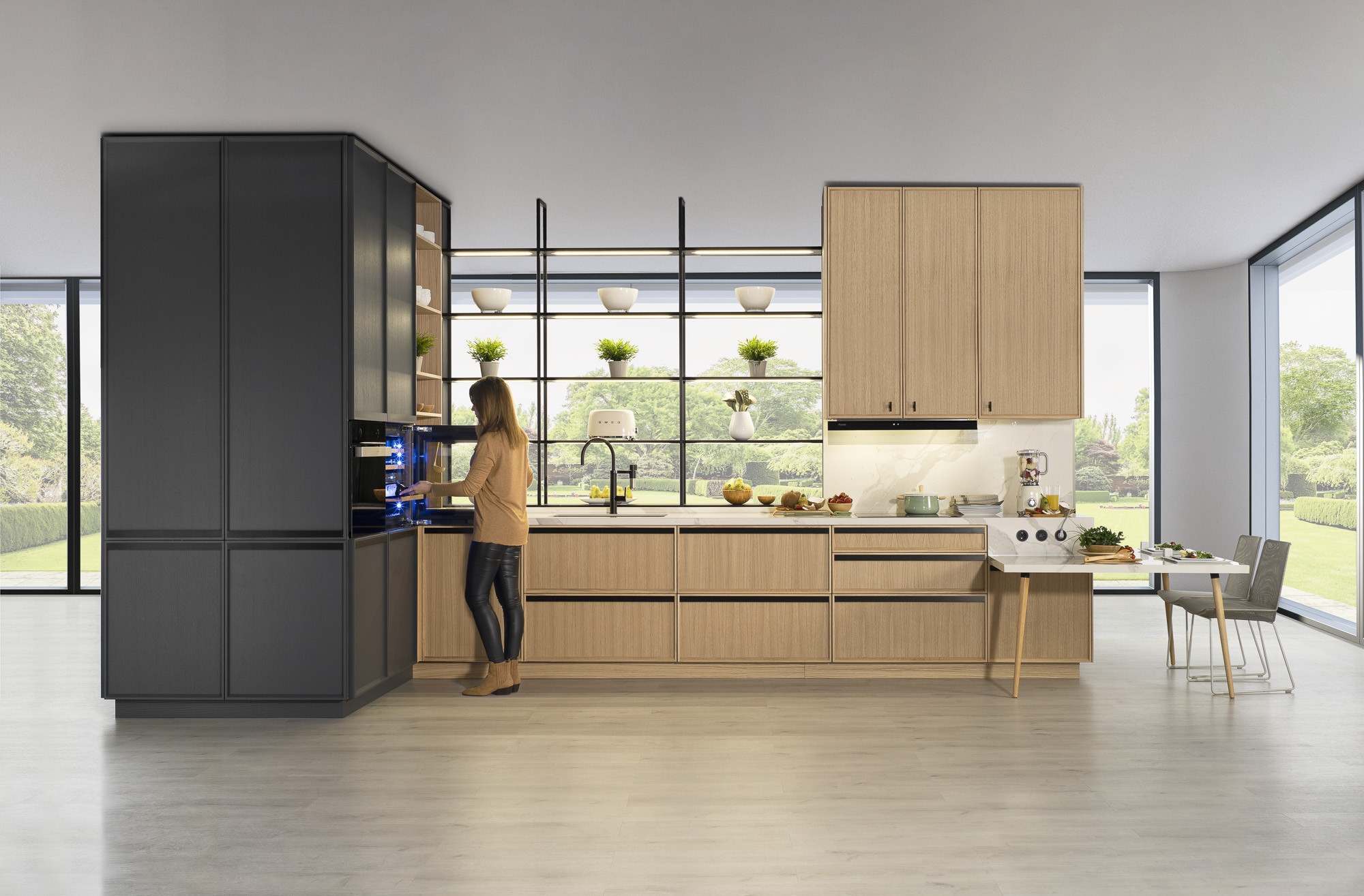 Cocina de estilo moderno con muebles de líneas limpias en acabado madera clara. Un módulo alto negro alberga electrodomésticos integrados, mientras que la isla se extiende con una mesa auxiliar tipo barra. Estanterías abiertas frente a ventanales amplios aportan luz natural y un toque decorativo con plantas y vajilla.