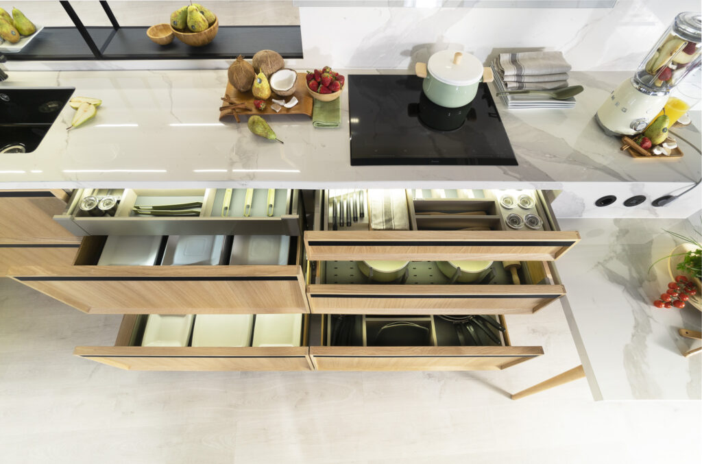Vista superior de una cocina con encimera de mármol claro y mobiliario en madera clara. Los cajones están abiertos, mostrando una organización precisa: cubertería, vajilla, utensilios y especieros distribuidos en compartimentos internos iluminados. Destaca la placa de inducción empotrada y la cuidada disposición de alimentos y utensilios sobre la encimera.