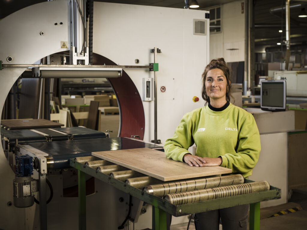 Una trabajadora posa con confianza frente a una máquina industrial, mientras sostiene una tabla de madera sobre la cinta transportadora. El entorno transmite orden, precisión y experiencia en procesos de manufactura.
