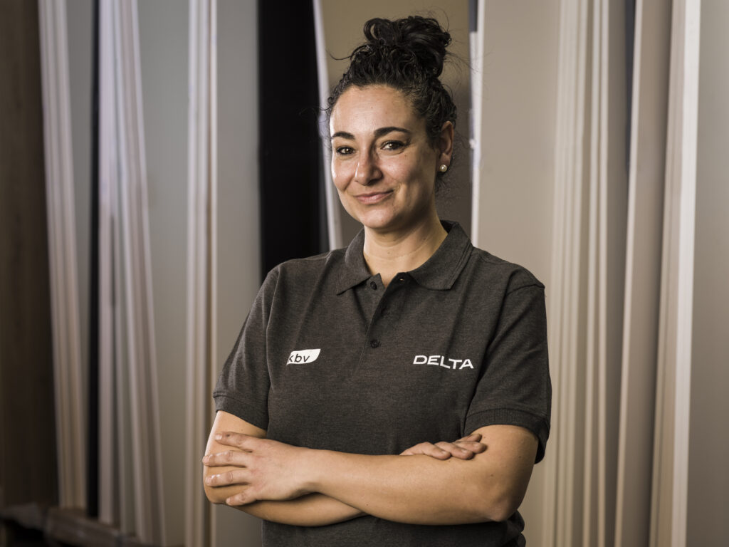 Esta imagen muestra a una mujer con expresión segura, de pie con los brazos cruzados, vistiendo un uniforme gris con el logo de "DELTA" y "kbv". El fondo está compuesto por paneles verticales que podrían ser muestras de materiales o puertas de muebles, lo cual sugiere un entorno relacionado con la fabricación o diseño de interiores.
