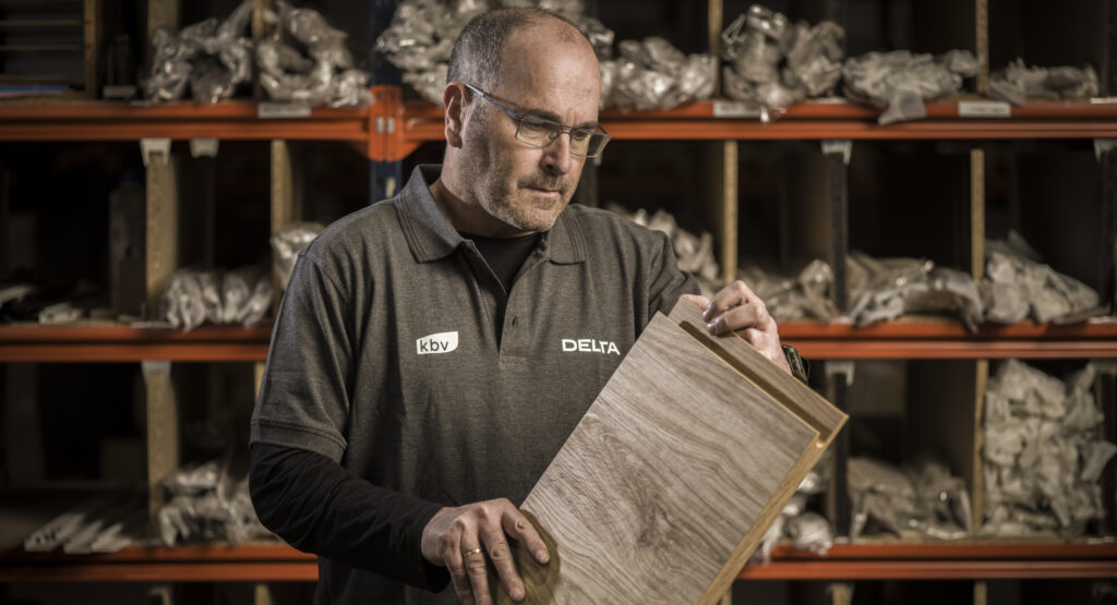 Un profesional examina con atención una muestra de madera, evaluando su textura y acabado. Al fondo, estanterías con piezas ordenadas sugieren un entorno de trabajo técnico y meticuloso.