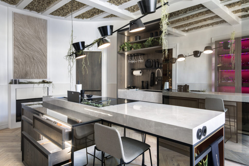 Una cocina moderna y elegante con una gran isla central de mármol claro. Los cajones están abiertos mostrando soluciones de almacenamiento funcional. Hay lámparas colgantes negras que añaden un toque sofisticado, y vegetación decorativa colgando del techo. En el fondo se ve una zona de copas y utensilios de cocina bien organizados.