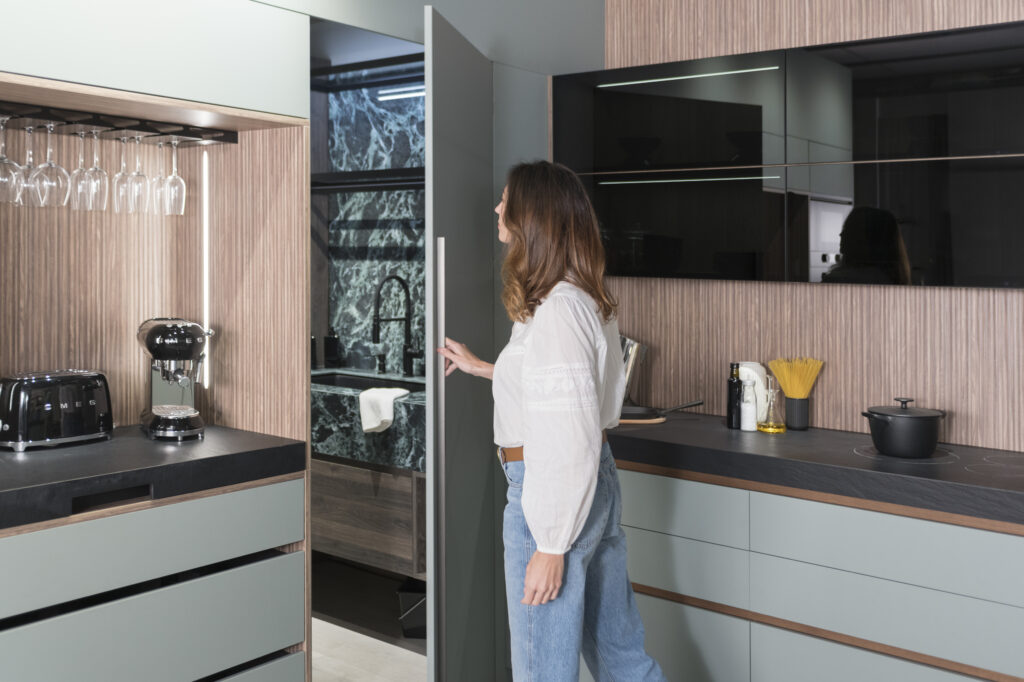 Puerta oculta integrada en el mobiliario de cocina, que conduce a un espacio adicional con acabados en mármol oscuro y grifería negra. La continuidad estética se mantiene gracias a los frentes de mueble en tono verde suave y sin tiradores visibles, acentuando una estética minimalista y funcional.