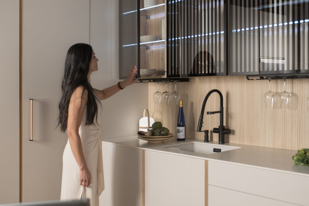 Elegancia minimalista con iluminación tenue. Puertas de vidrio con lamas oscuras revelan vajilla ordenada. Grifería negra y detalles en madera completan un rincón de calma y estilo.