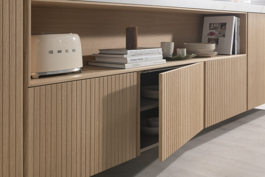 Frentes acanalados en madera clara que ocultan espacio de almacenaje, combinados con un estante abierto para mostrar libros y vajilla. Detalles minimalistas resaltan la textura natural y aportan calidez al diseño.