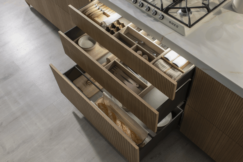 Cajonera de isla con frentes acanalados en madera clara, tres gavetas de gran profundidad equipadas con divisores modulares de madera para cubiertos, utensilios y pan. Organización inteligente que combina estética y practicidad.