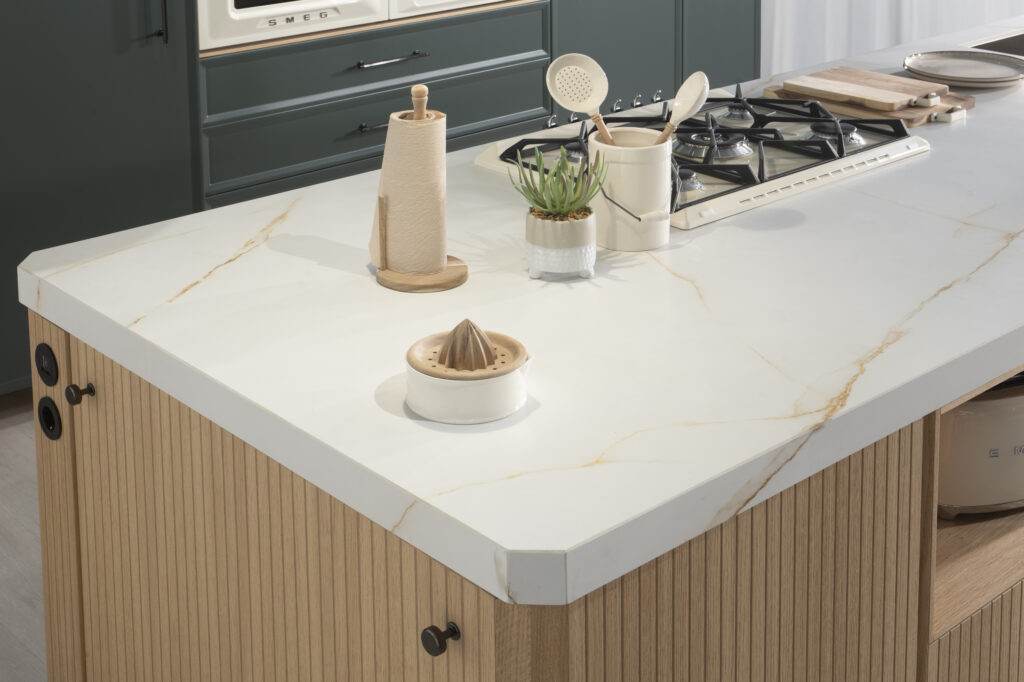Encimera blanca de efecto mármol con sutiles vetas doradas, instalada sobre una isla de frentes acanalados en madera clara. Placa de gas integrada y accesorios minimalistas (porta rollos, utensilios, planta) realzan su funcionalidad y elegancia.