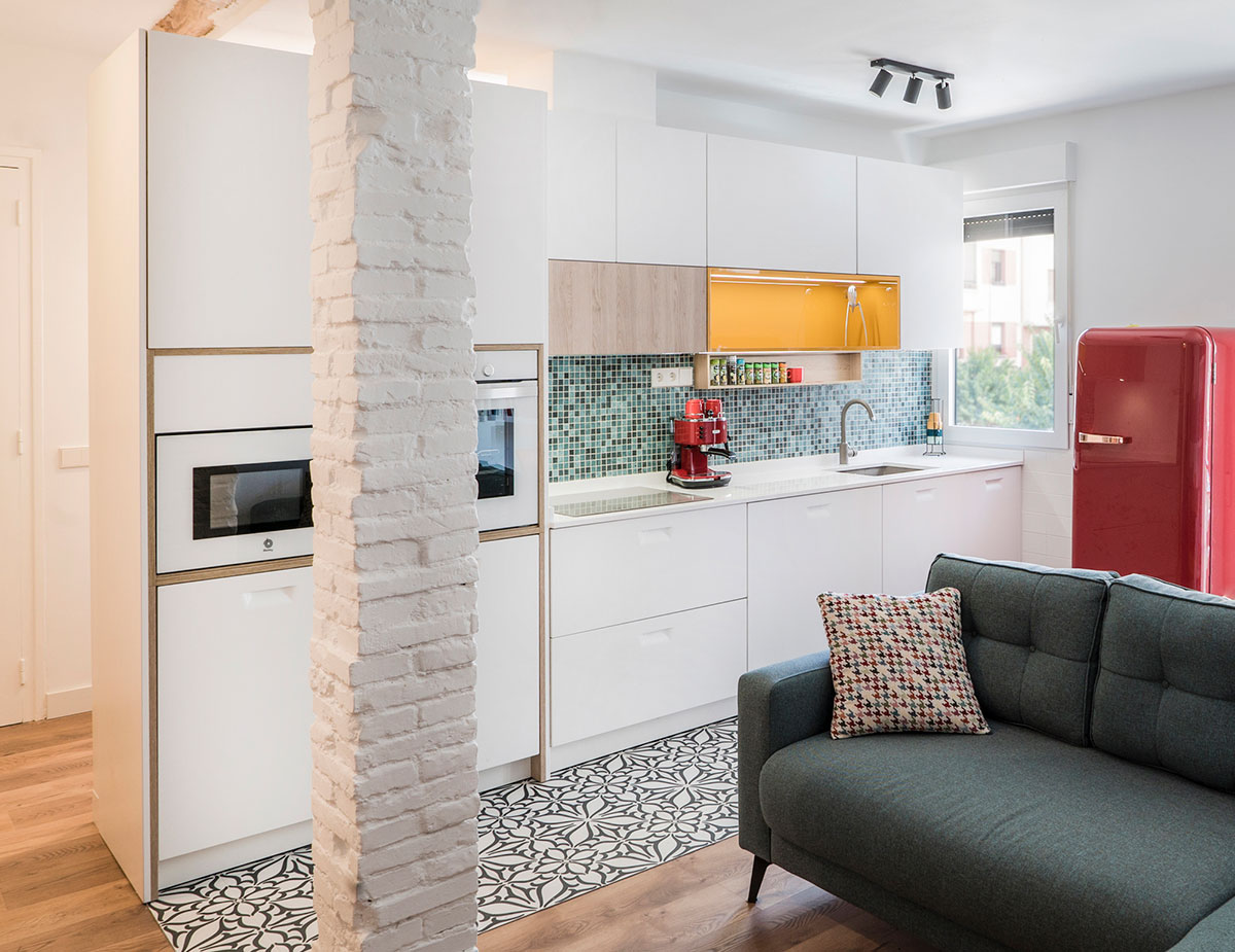Cocina compacta y luminosa con muebles blancos de líneas limpias, realzada por detalles en madera clara y un toque de color mostaza en los módulos superiores. El salpicadero de mosaico y la nevera retro en rojo aportan personalidad y frescura al espacio.